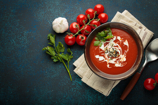 Fresh tomato soup is served in a bowl with cream, seeds and herbs, accompanied by cherry tomatoes, garlic and parsley on a blue background with a spoon and napkin, copy space