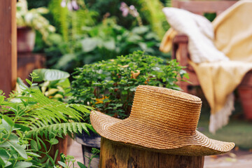 Straw hat placed on tree stump surrounded by green foliage and potted plants. Wooden armchair with soft throw sits blurred in background, evoking warm, homely garden vibe for relax in summer day