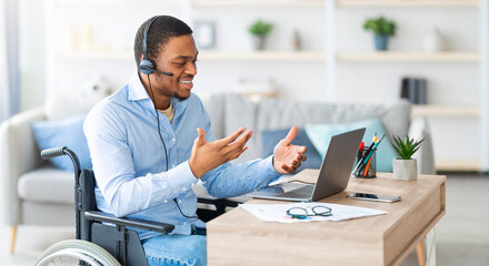 Black guy with disability with headset communicating online on laptop, having business meeting from home. Millennial African American man in wheelchair working in call center remotely