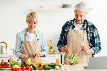 Couple preparing healthy vegetarian salad, enjoying process on modern cozy kitchen, cooking together, relish activity at home. Smiling adult lady and man in aprons at table with vegetables, copy space