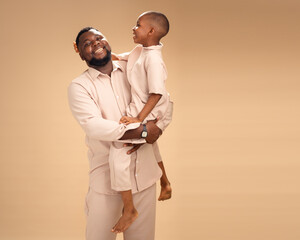 In warm studio light, an African father embraces his young son in matching beige. Their close pose...