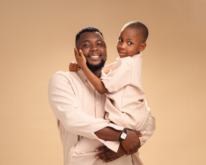A joyful African father embraces his son, both facing the camera in matching beige outfits. Soft...