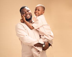 A happy African father holds his smiling son in a warm embrace. Both wear matching beige outfits in...