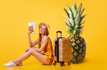 A young woman in a vibrant yellow outfit sits with a suitcase and pineapple, holding travel...