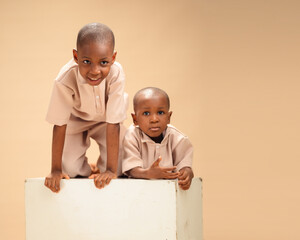 Two young Black brothers in matching beige outfits, posing playfully on a white block in indoor...
