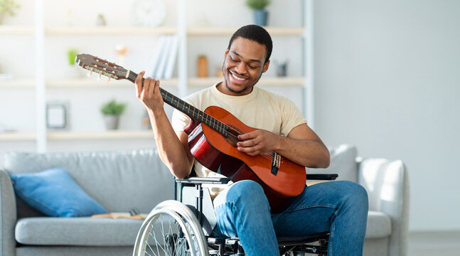 Black guy in wheelchair playing guitar and smiling at living room. African American man with disability with instrument performing beautiful music indoors. Stay home activities