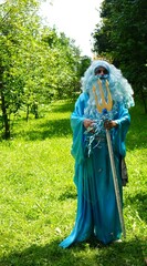  Person dressed as Poseidon in a garden setting, holding a trident, with blue wig and robe, evoking a mythical and theatrical atmosphere in bright daylight.