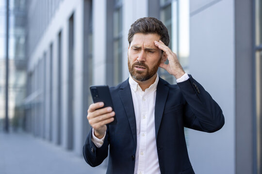 A businessman in a suit appears stressed while holding and using a smartphone outdoors. The scene conveys emotions of confusion or anxiety, set in a modern urban environment during daylight.