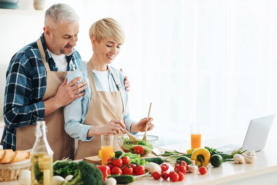 Senior couple bloggers recording video content, preparing healthy food in kitchen. Caucasian man hugs smiling wife making salad in kitchen interior with bright fresh vegetables on table with laptop - Powered by Adobe