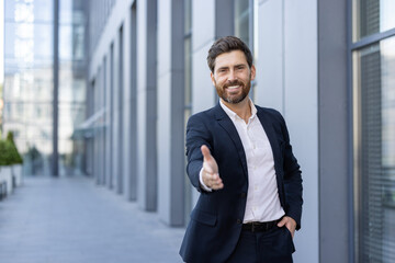 Confident male professional extending a handshake outdoors in a corporate office setting,...