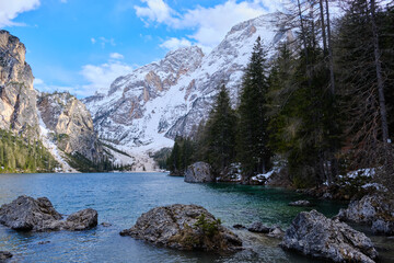 March 2025: view of Braies Lake in winter, with a frozen surface marked by trails, surrounded by snow covered cliffs, rugged rocks, and evergreen trees.a