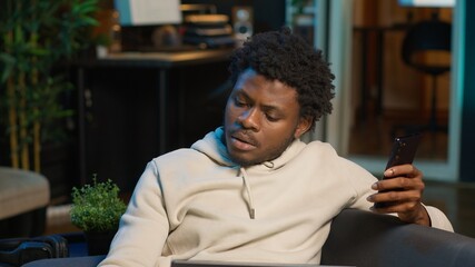 Black freelancer does his research by reading articles and data driven papers at home, using phone...