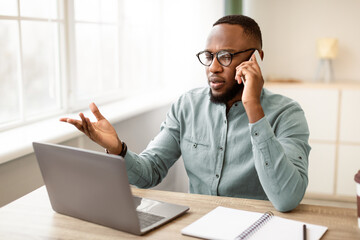 Serious Black Businessman Talking On Mobile Phone At Laptop Computer At Workplace Sitting At Desk...