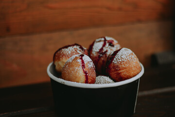 Delicious round donuts with powdered sugar and berry sauce served in a black paper cup. Rustic wooden table with yellow chrysanthemums in the background. Cozy autumn food scene.