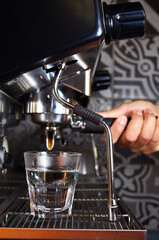 Side view of a barista operating a professional espresso machine, preparing a drink with a transparent glass placed under the group head. The machine's steam wand and the barista&rsquo;s hand are visible