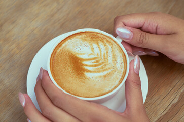 Top view of a beautifully crafted latte with foam art in a white ceramic cup, gently held by a person with manicured hands on a wooden table.