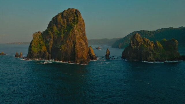 Aerial view of massive coastal rock formations rising from deep waters with forested hills behind
