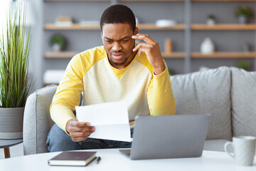 Confused black man sitting on couch in front of laptop, holding papers and touching his head, home...