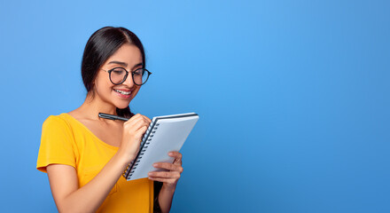 Fototapeta premium Checklist. Portrait of intelligent indian female student wearing glasses, taking notes in copybook on purple studio background. Smart young woman studying, preparing for exam, writing in notebook