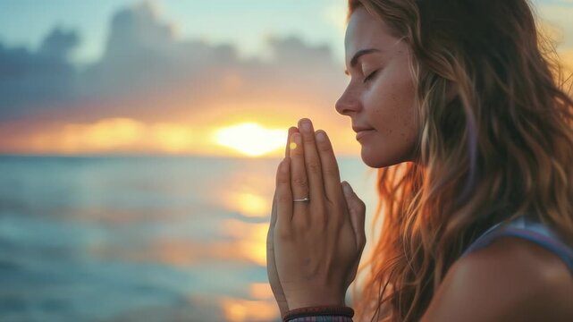 Close-up woman meditating on beach during sunset. She is holding her hands together in prayer position, her eyes are closed, sun is setting over ocean in background.