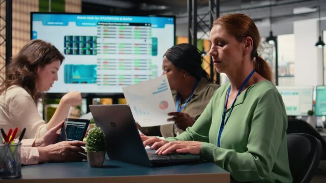 Determined female CEO reviews graphs and performance reports on her laptop, meeting with female managers in a modern corporate office to ensure commitment to business growth. Camera A.