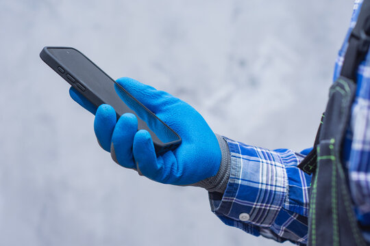 Close-up of a construction worker handyman in protective gloves and workwear using a modern smartphone. Concept digital technology into manual labor and job.