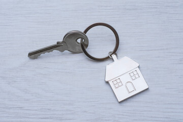 Metal house key attached to silver keyring with house-shaped keychain on white wooden surface. Real estate and property ownership concept.