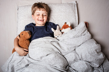 Happy child waking up in bed with big smile and raised arm, surrounded by stuffed animals. Joyful...