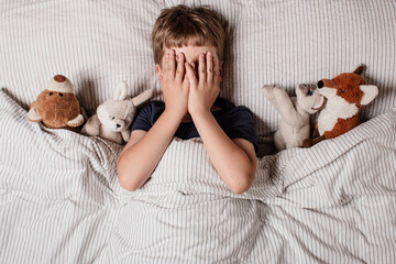 The boy was scared before going to bed. Night terrors in a child. The kid covers his face with his hands in a fear. Children's experiences. Boy in the bed. View from above. Sad psychological state.