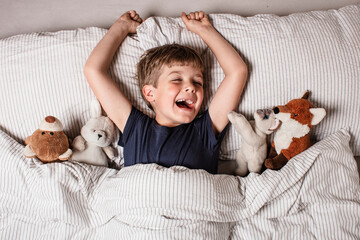 Happy child waking up in bed with big smile and raised arm, surrounded by stuffed animals. Joyful...