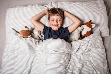 Happy child waking up in bed with big smile and raised arm, surrounded by stuffed animals. Joyful...