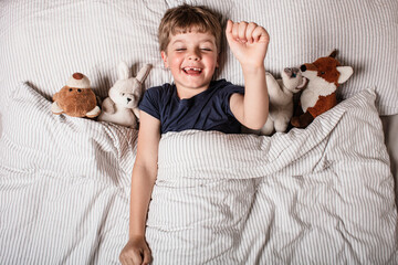 Happy child waking up in bed with big smile and raised arm, surrounded by stuffed animals. Joyful...