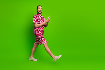 Cheerful Young Man in Pink Leopard Print Outfit Using Smartphone Against Solid Green Background,...