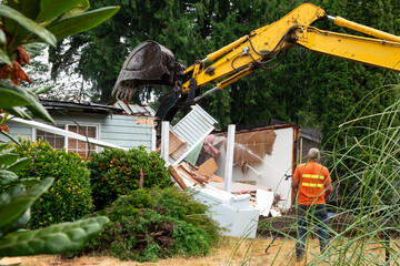 Workman with hose spraying water on demolition of old wood frame house for redevelopment of...