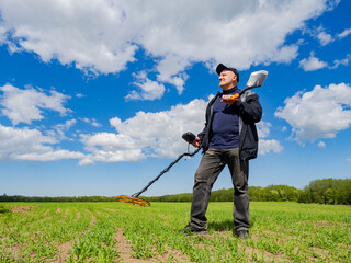 Man with metal detector and shovel exploring green field under blue sky, searching for treasures or...