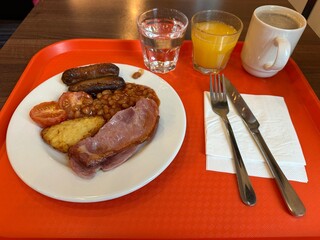 Full English breakfast plate including bacon, potato toastie, tomato, sausages, and beans with coffee, juice, and water to drink on an orange tray