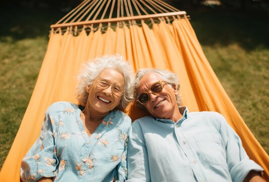 An affectionate elderly couple relaxes in a hammock, surrounded by lush greenery. They smile joyfully at the camera, dressed casually in light clothing and sunglasses.