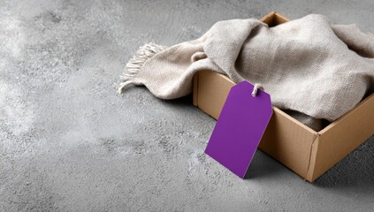 A light beige/taupe scarf draped over the open top of a cardboard box, with a purple price tag attached.  The box and scarf rest on a speckled gray concrete surface
