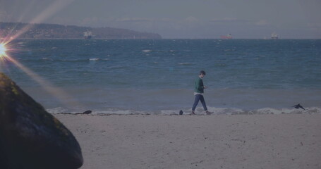 Walking man holding smartphone on sandy beach, with ocean waves, mossy rock, seagulls, cargo ships