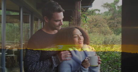 Cuddling couple wearing knit sweaters on veranda railing, gazing at hedgerow holding white mug