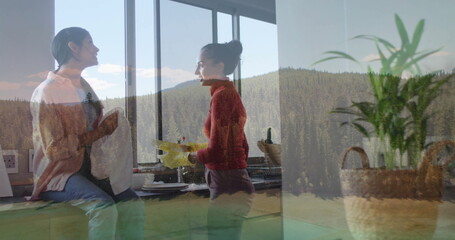 Mother and daughter chatting while holding mug and washing dishes wearing yellow gloves in kitchen