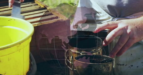 Operating beekeeper in grey zip-up top using bee smoker and hive tool at apiary with bucket