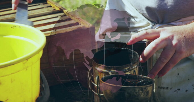 Operating beekeeper in grey zip-up top using bee smoker and hive tool at apiary with bucket