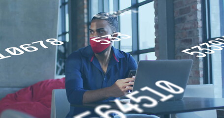 Analyzing man in red mask using laptop and smartphone in coworking lounge with numerical overlays
