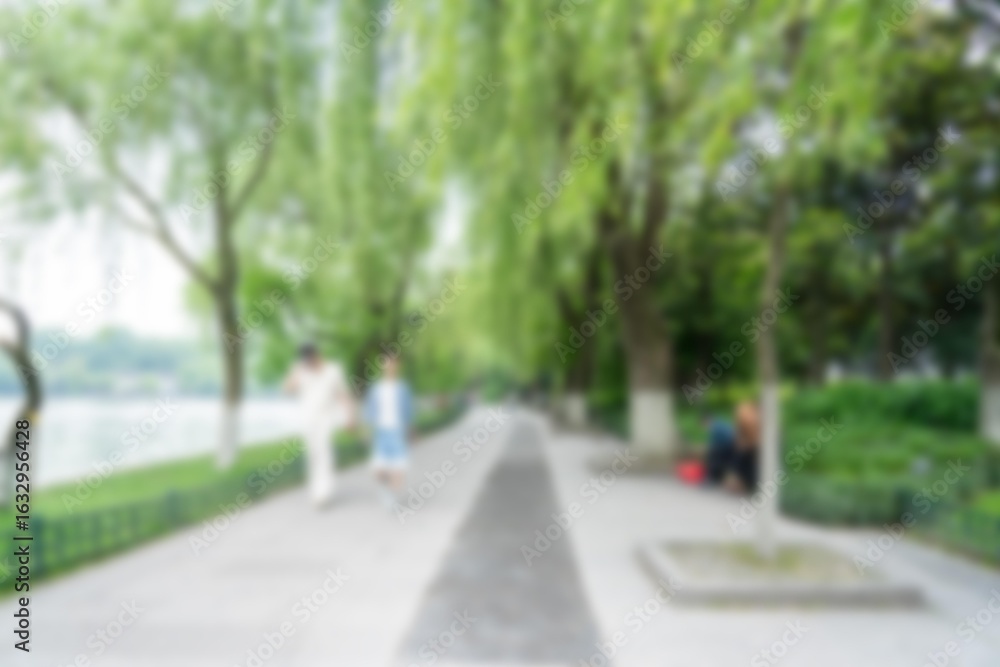 Poster a blurred image of a walkway lined with trees and greenery, with a glimpse of a body of water in the