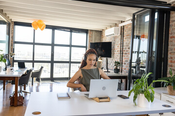 African American woman typing on laptop at office desk with notebook, smartphone and headphones