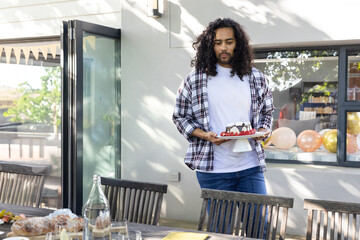 Middle Eastern male holding celebration cake near dining table with balloons in sunlight on patio