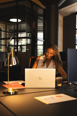 Woman wearing sleeveless blouse working on laptop at desk in modern office with lamp, copy space