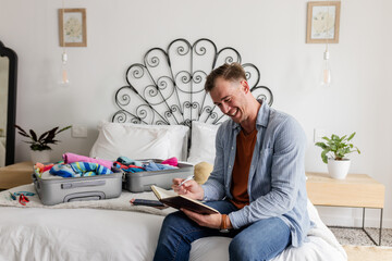 Man sitting on bed writing in notebook with open suitcase in bedroom, copy space