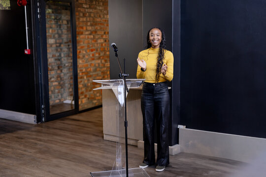African American woman in mustard top clapping behind clear lectern with microphone in event space